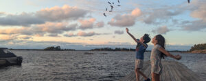 Two figures on a weathered wooden dock, silhouetted against an orange-pink sky. They playfully toss small objects into the a