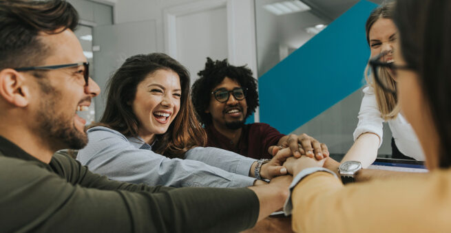 People in an office placing their hands together, symbolizing teamwork and unity.